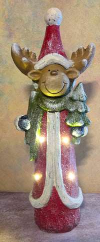 Front view of tall light-up reindeer Christmas figurine wearing a red coat, green scarf, oversized Santa hat, and holding a snowy evergreen tree.
