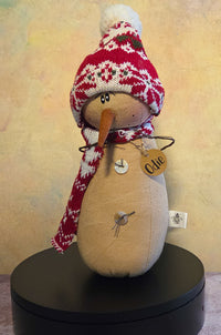 Odie the Snowman winter soft sculpture shelf sitter with red knit hat, matching scarf, carrot nose, twig arms, button details, and name tag.