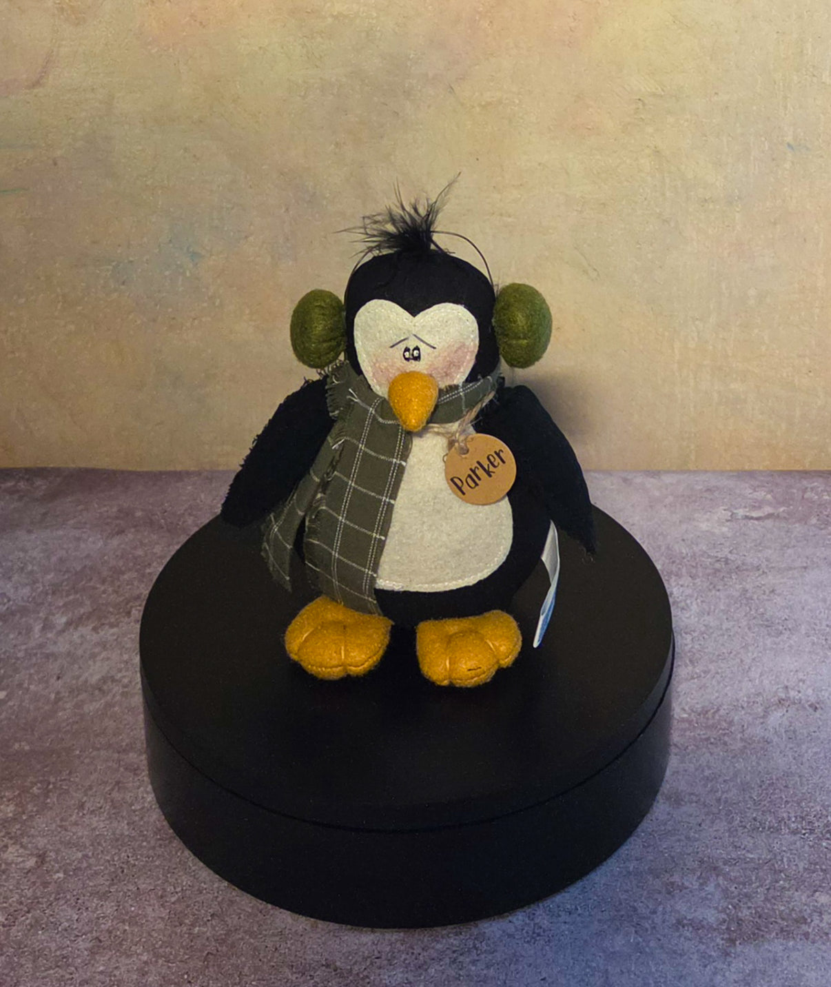 Front view of Parker penguin soft sculpture with green earmuffs, plaid scarf, golden feet, and name tag.