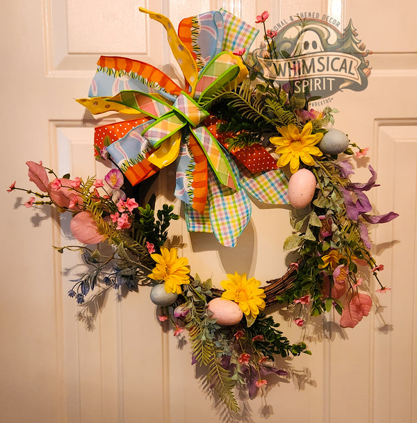 Decorative wreath with colorful flowers and a large bow on a door, featuring the brand 'Whimsical Spirit'.