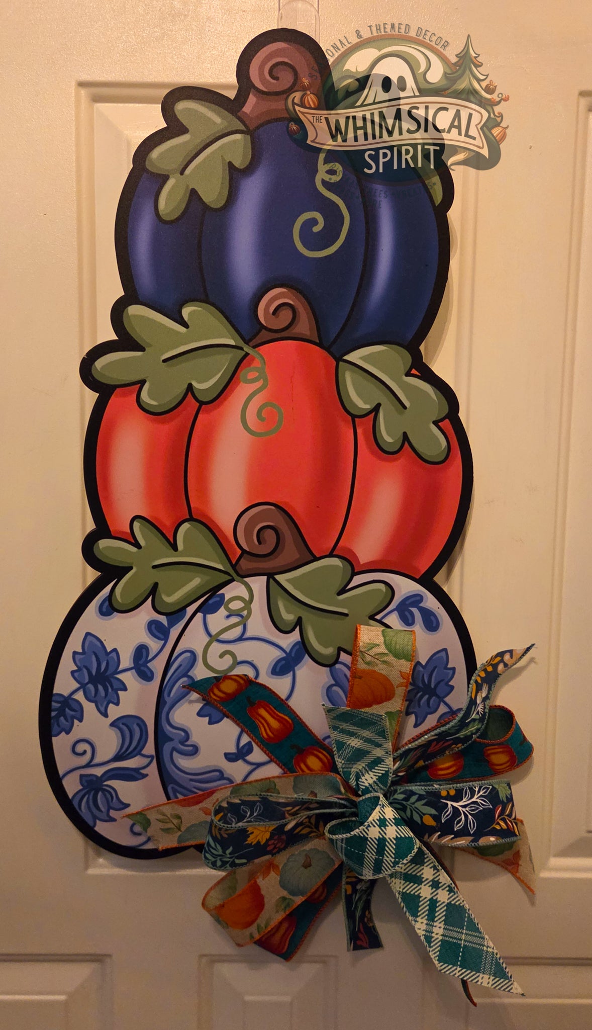 Stacked fall pumpkin door hanger featuring a navy pumpkin, coral-orange pumpkin, and blue-and-white floral pumpkin with green leaves and a layered teal, navy, and fall-patterned bow.
