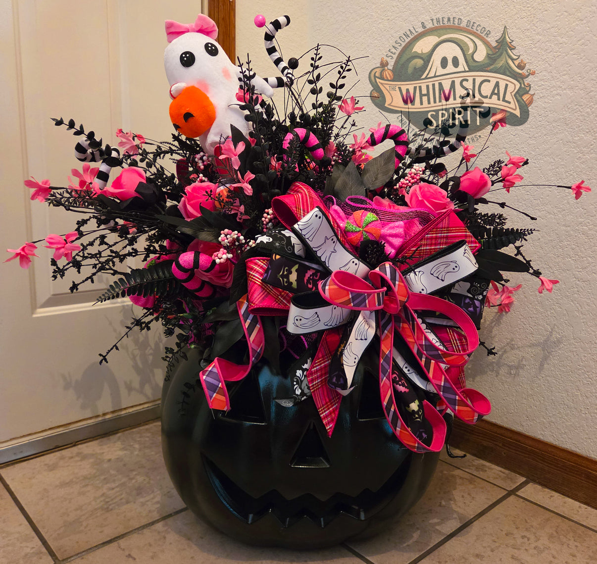 Front view of an oversized black jack-o’-lantern decorated with pink florals, black foliage, striped accents, a white ghost, and layered Halloween ribbons.