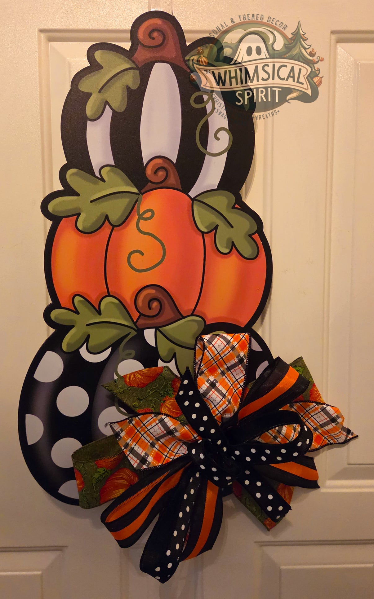 Stacked fall pumpkin door hanger hanging on a white paneled door, featuring black-and-white striped and polka dot pumpkins, green leaf accents, and a large black, orange, plaid, and polka dot bow at the bottom.