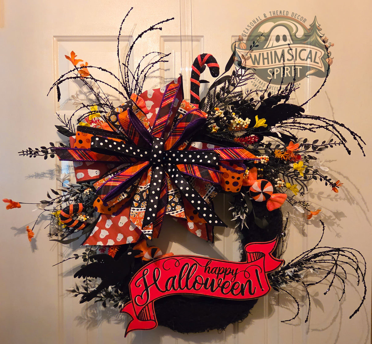 Handmade oval moss Halloween wreath with black ravens, a Happy Halloween banner, curled black sprays, candy accents, and a large patterned bow.