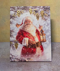 Front view of the light-up Christmas tabletop canvas featuring Santa holding a lantern beneath snowy evergreen branches and pinecones.