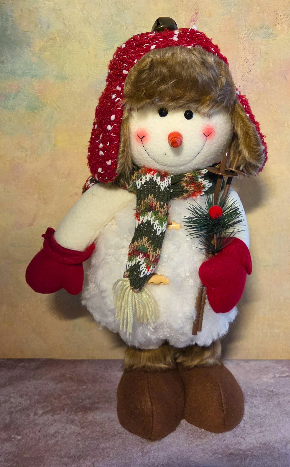 Tall skier snowman soft sculpture displayed with its adjustable legs shortened, wearing a fur-trimmed hat, patterned scarf, and red mittens.