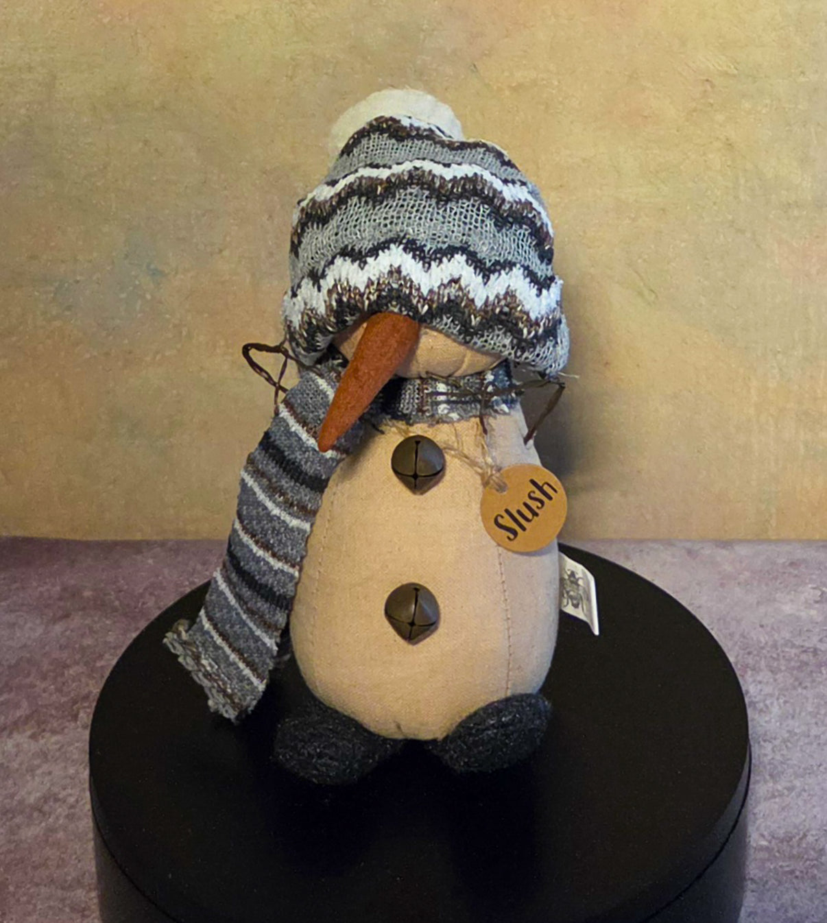 Front view of Slush snowman soft sculpture with a striped knit hat and scarf, long carrot nose, jingle bell buttons, and wooden name tag.