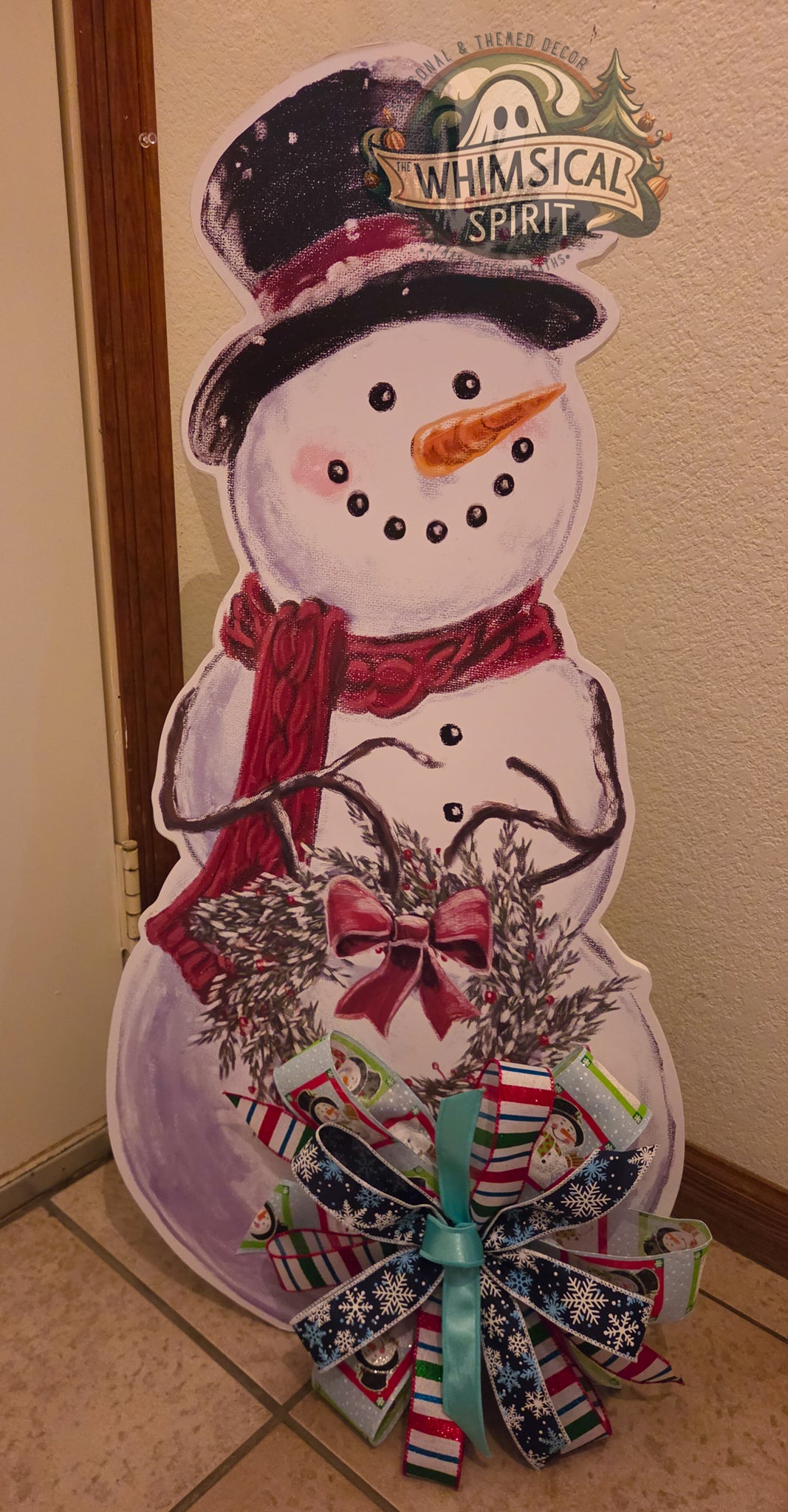 Snow Much Cheer freestanding snowman porch sitter with a black top hat, red scarf, evergreen wreath, and colorful Christmas bow.