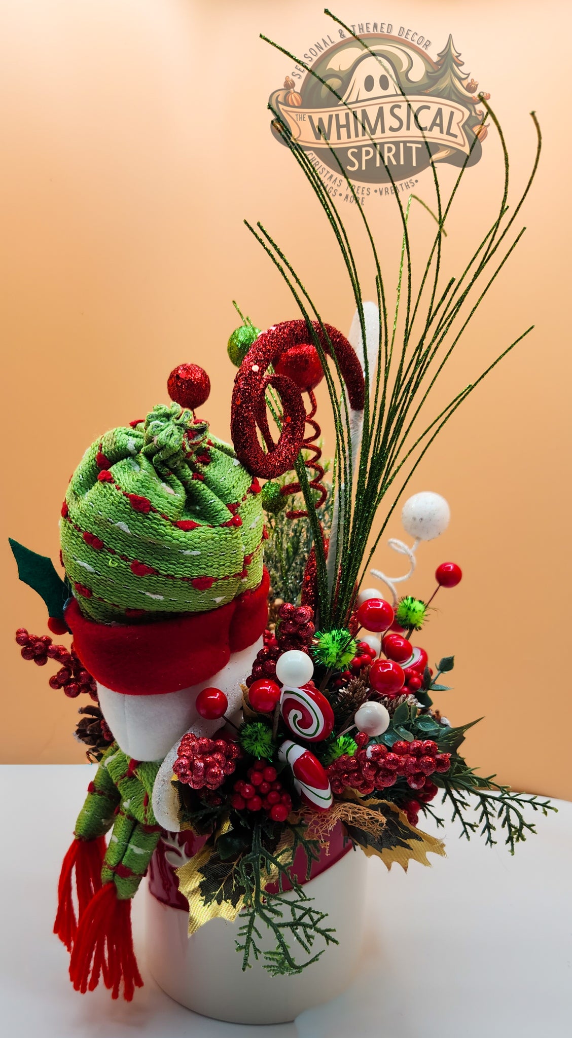 Decorative Christmas arrangement with a green hat, red scarf, and candy cane in a vase on a beige background.