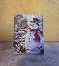 Snowman and Feathered Friend light-up Christmas tabletop canvas featuring a smiling snowman, red cardinal, snowy cottage, berries, and winter scene.