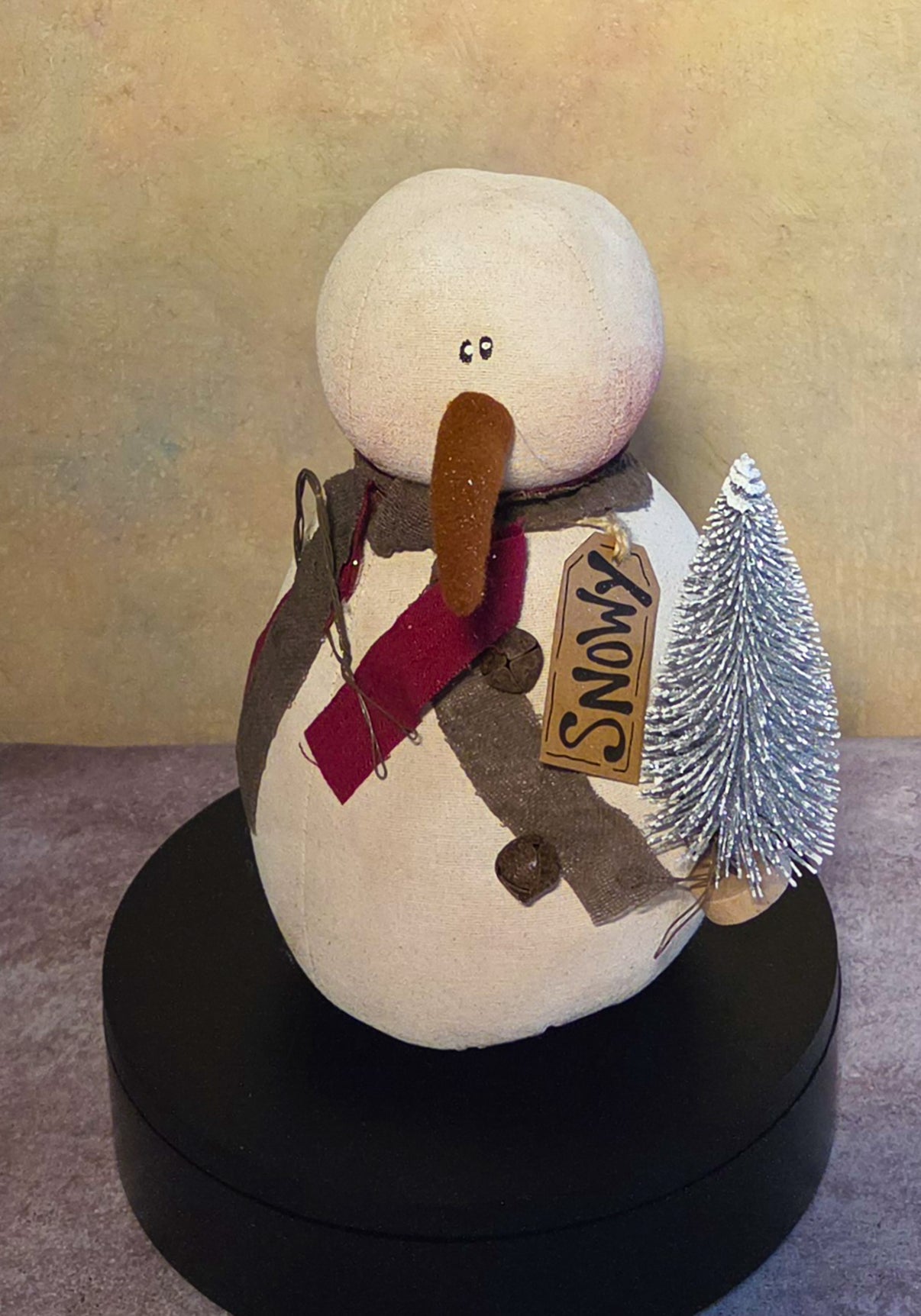 Front view of Snowy snowman soft sculpture with a brown carrot nose, burgundy and gray scarf, name tag, buttons, and frosted tree.