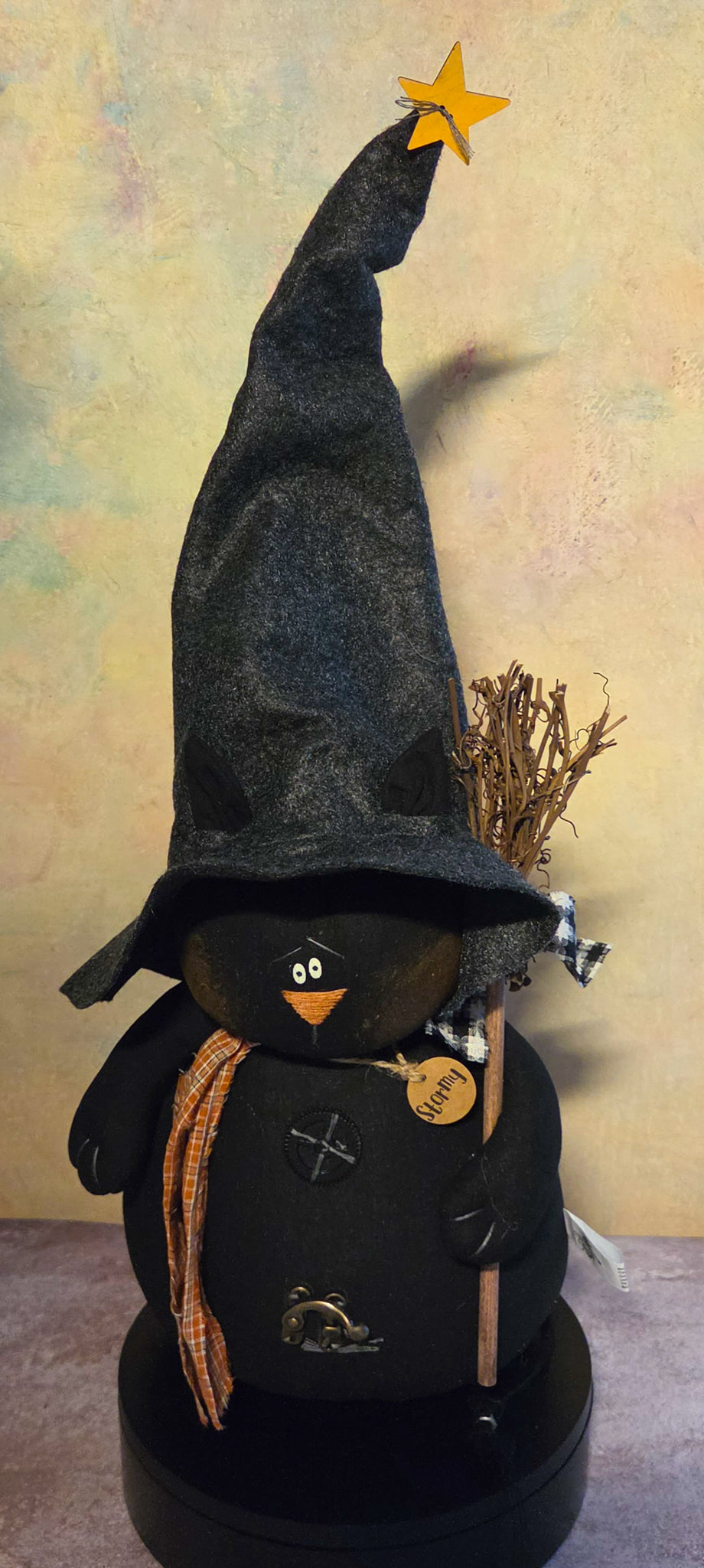 Front view of Stormy black cat soft sculpture with an extra-tall witch hat, orange plaid scarf, broom, rustic buttons, and wooden name tag.