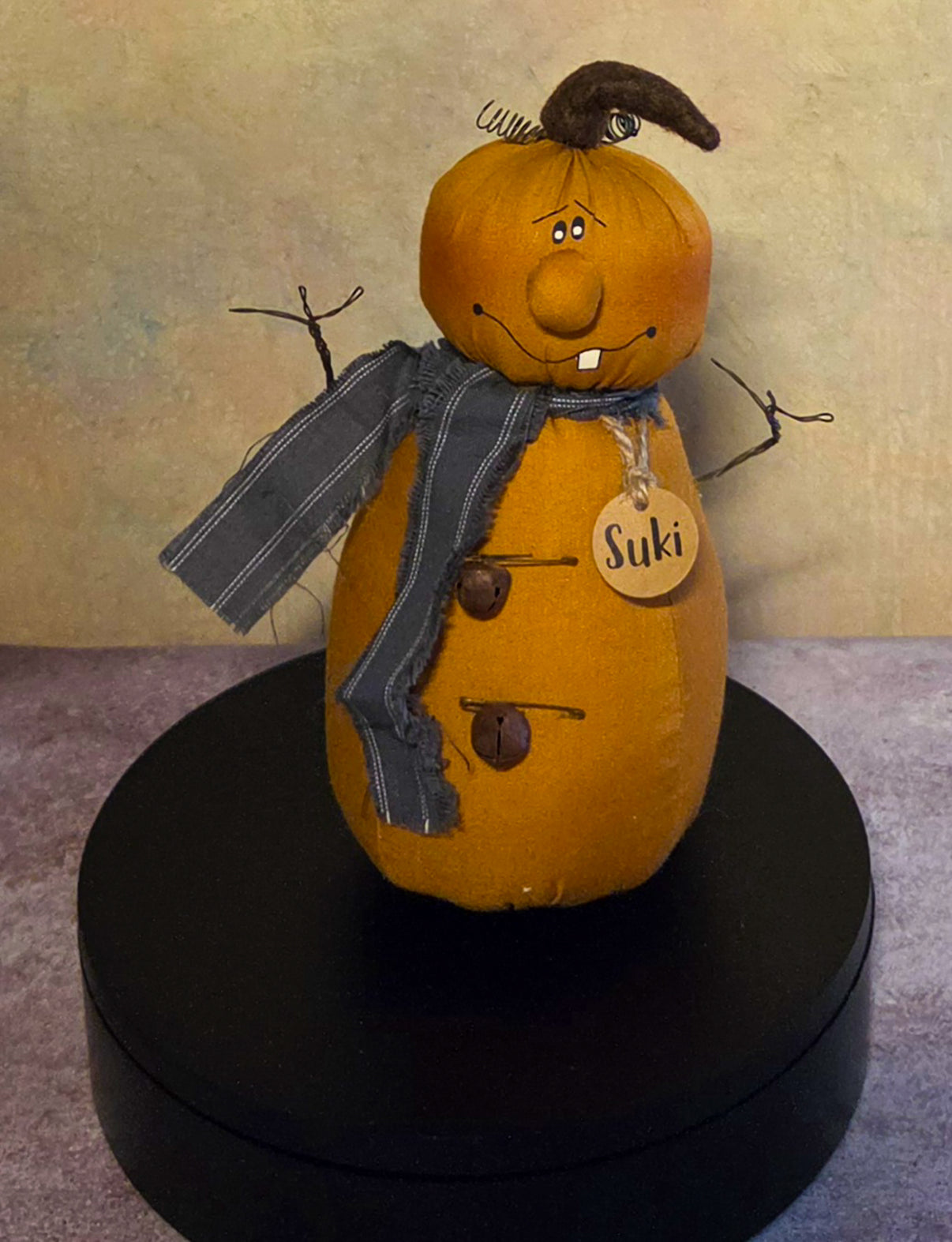 Front view of Suki jack-o’-lantern soft sculpture with a curved stem, twig arms, charcoal striped scarf, rustic bell buttons, and wooden name tag.