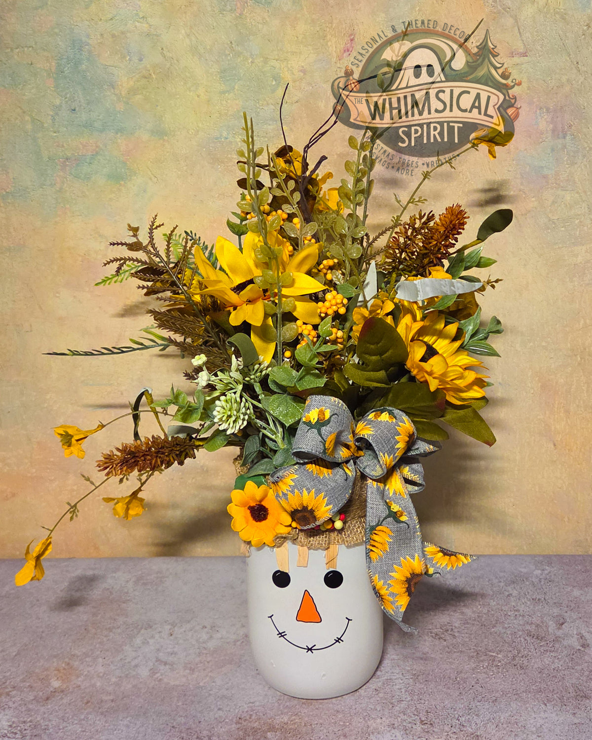 Tall fall centerpiece with a smiling white scarecrow container, yellow sunflowers, greenery, brown stems, and a gray sunflower bow.