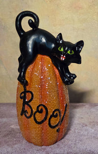 Crouching black cat with green eyes and a curled tail perched on a tall orange pumpkin labeled “Boo!”