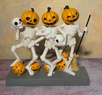 Three white skeletons with orange jack-o’-lantern heads, skulls, a broom, and small pumpkins displayed together on a gray base.