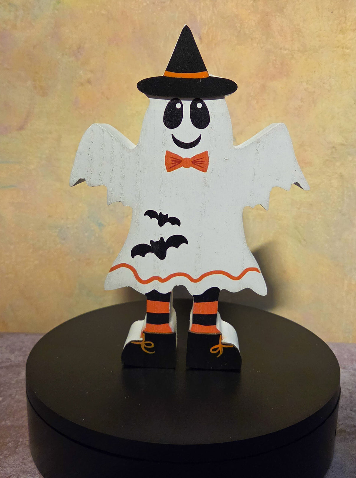 Front view of wooden Halloween ghost shelf sitter wearing a black witch hat, orange bow tie, striped legs, and bat accents.