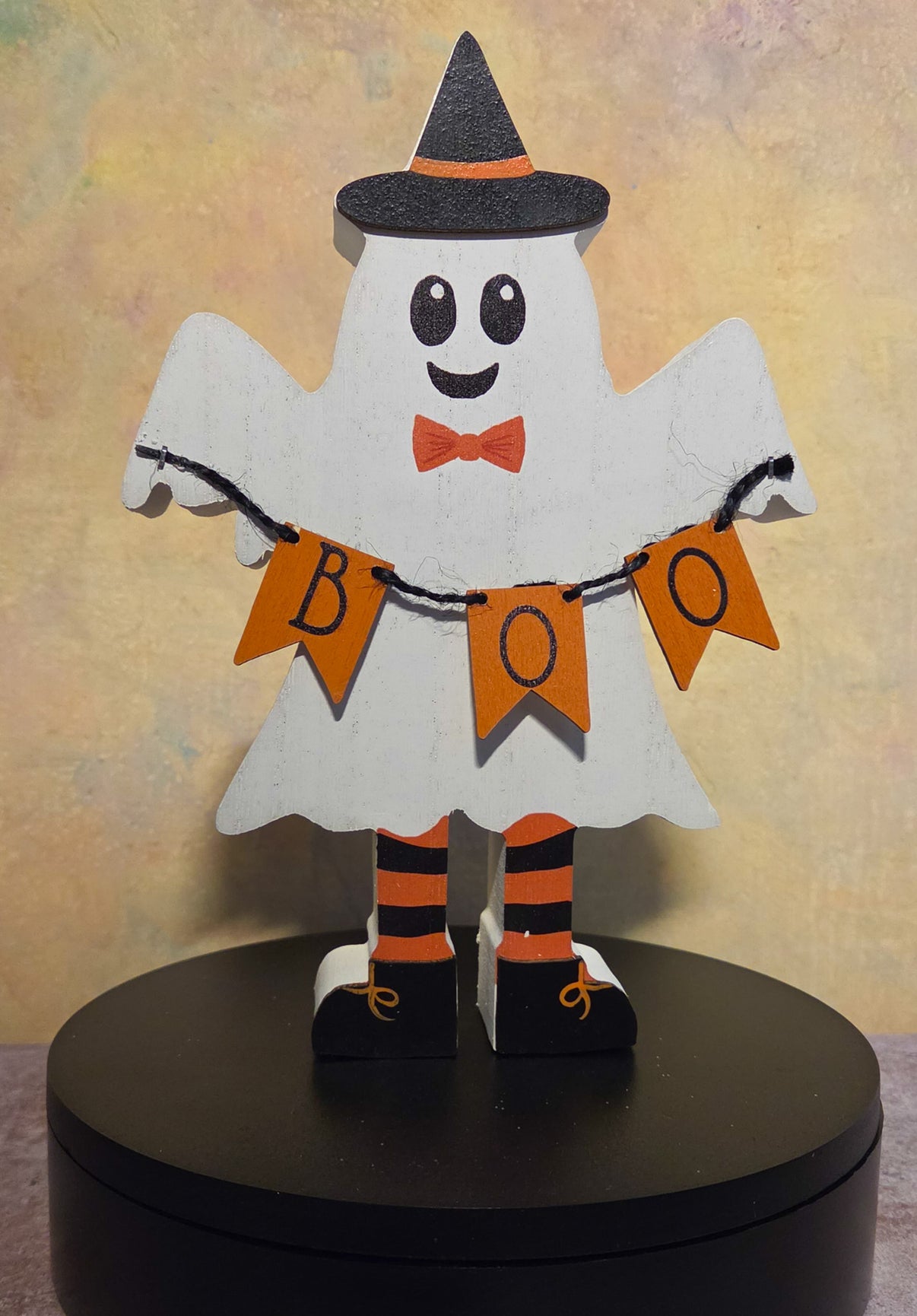 Front view of wooden Halloween ghost shelf sitter wearing a black witch hat and holding an orange BOO banner.