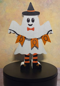 Front view of wooden Halloween ghost shelf sitter wearing a black witch hat and holding an orange BOO banner.