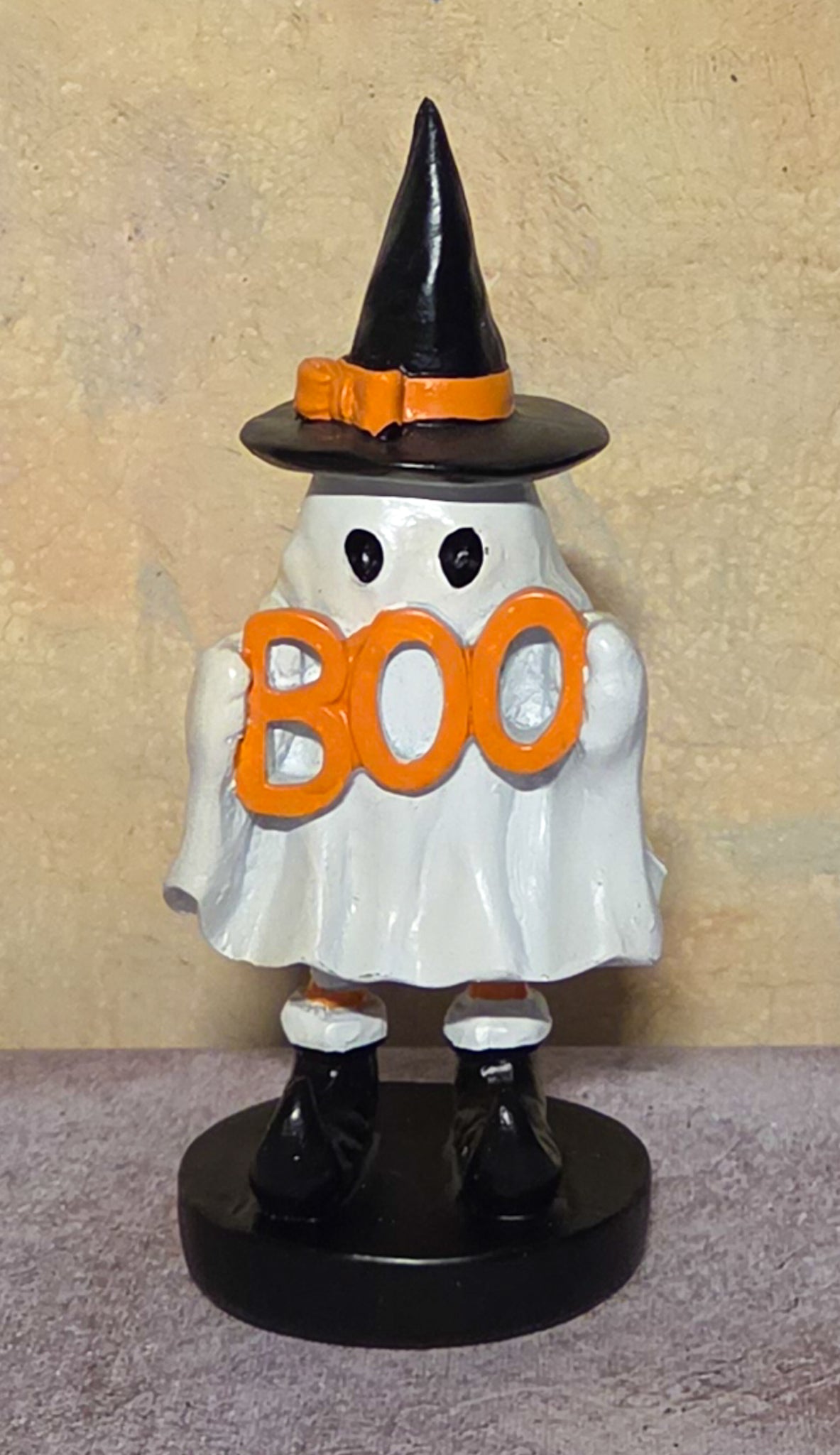 A Halloween ghost figurine wearing a black witch hat with an orange band, holding orange 'BOO' letters, standing on a black base against a textured beige wall.