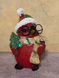 A red bird figurine wearing a white Santa hat with a pom-pom, round black glasses, a glittery gold scarf, and holding a small green tree, set against a textured beige background.