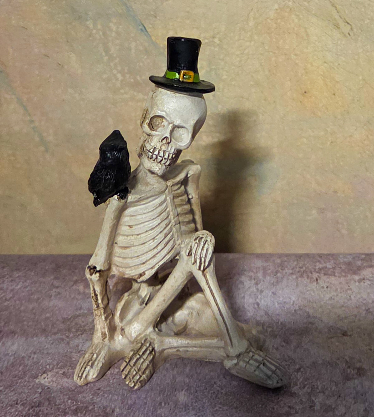 Seated skeleton figurine wearing a black top hat with a black raven perched beside its shoulder.