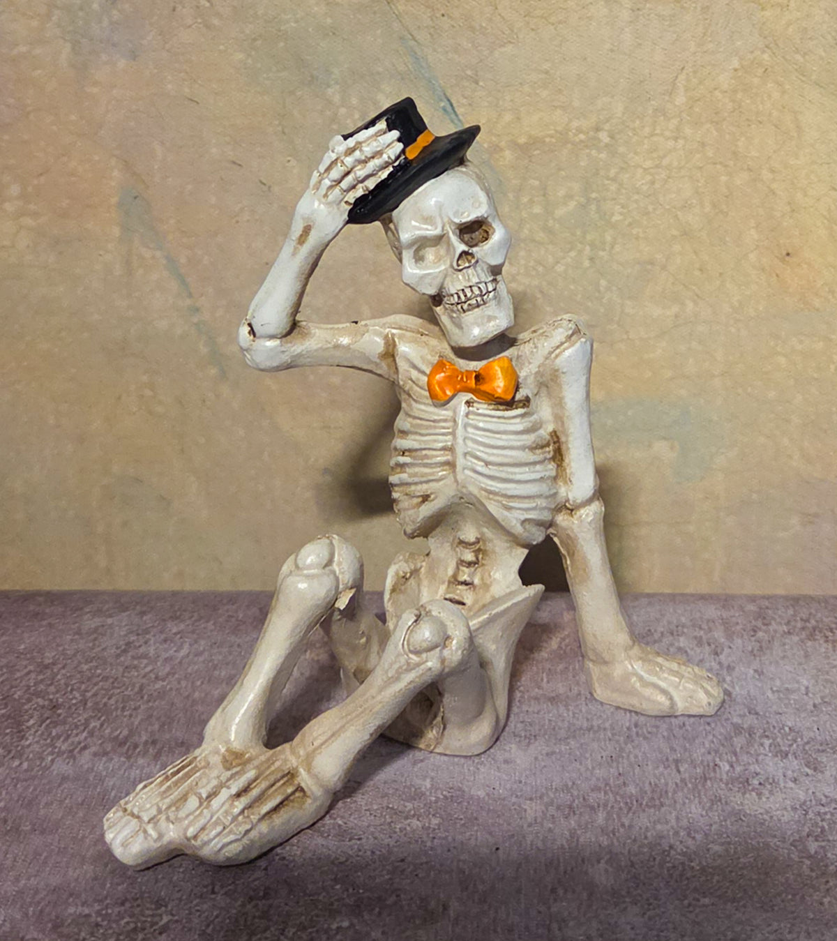 Seated skeleton figurine tipping a black top hat while wearing a bright orange bow tie.