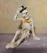 Seated skeleton figurine tipping a black top hat while wearing a bright orange bow tie.