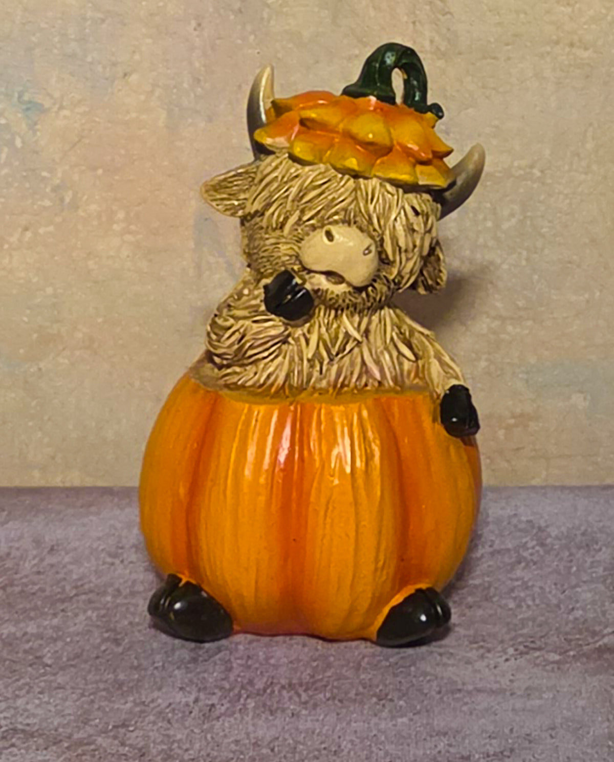 A decorative figurine of a shaggy Highland cow sitting inside a bright orange pumpkin, wearing a small orange and yellow pumpkin hat with a dark green stem, photographed against a textured beige wall and lavender surface.