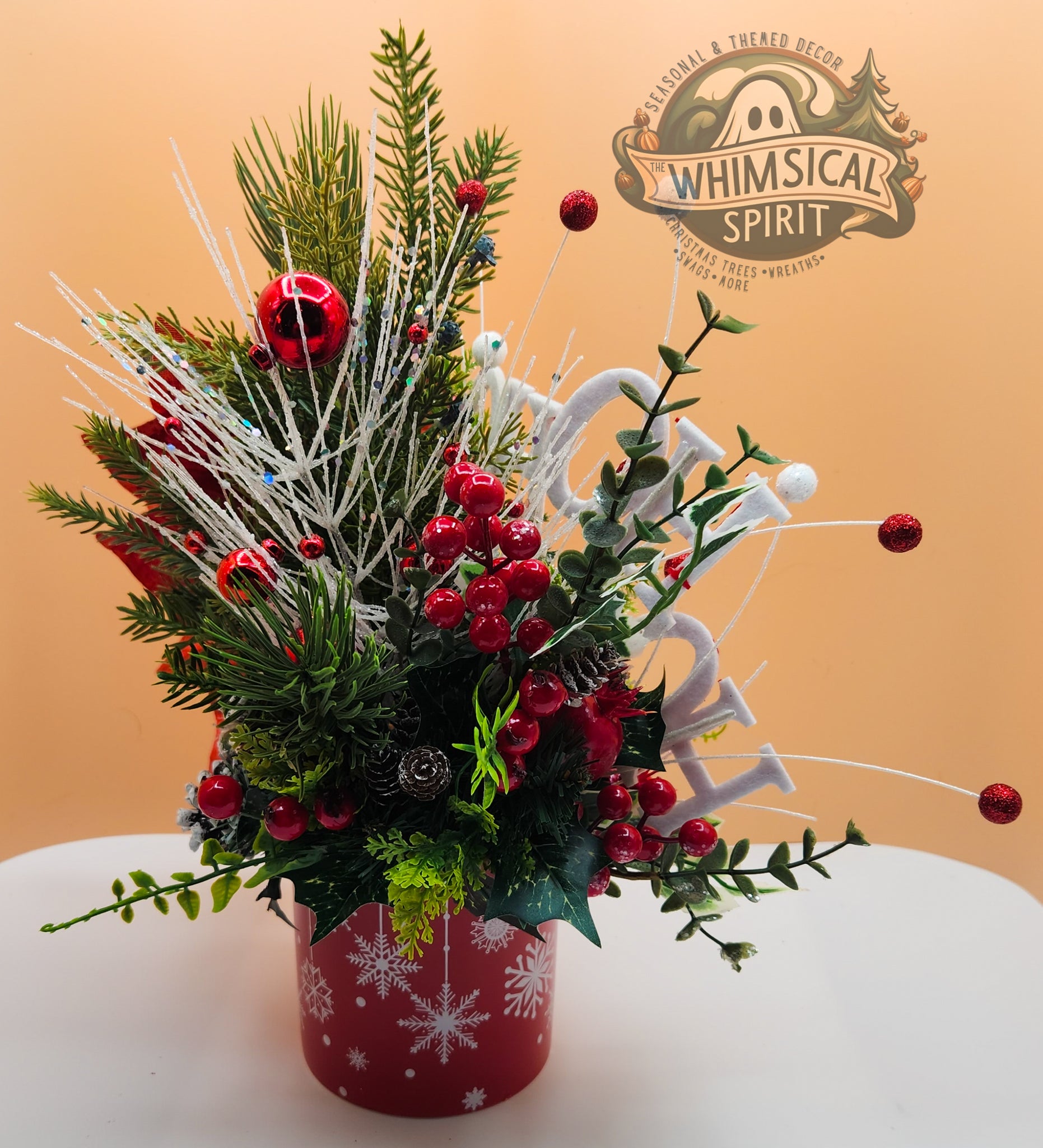 Christmas-themed floral arrangement with red berries and greenery in a red container on a beige background, featuring 'Whimsical Spirit' logo.