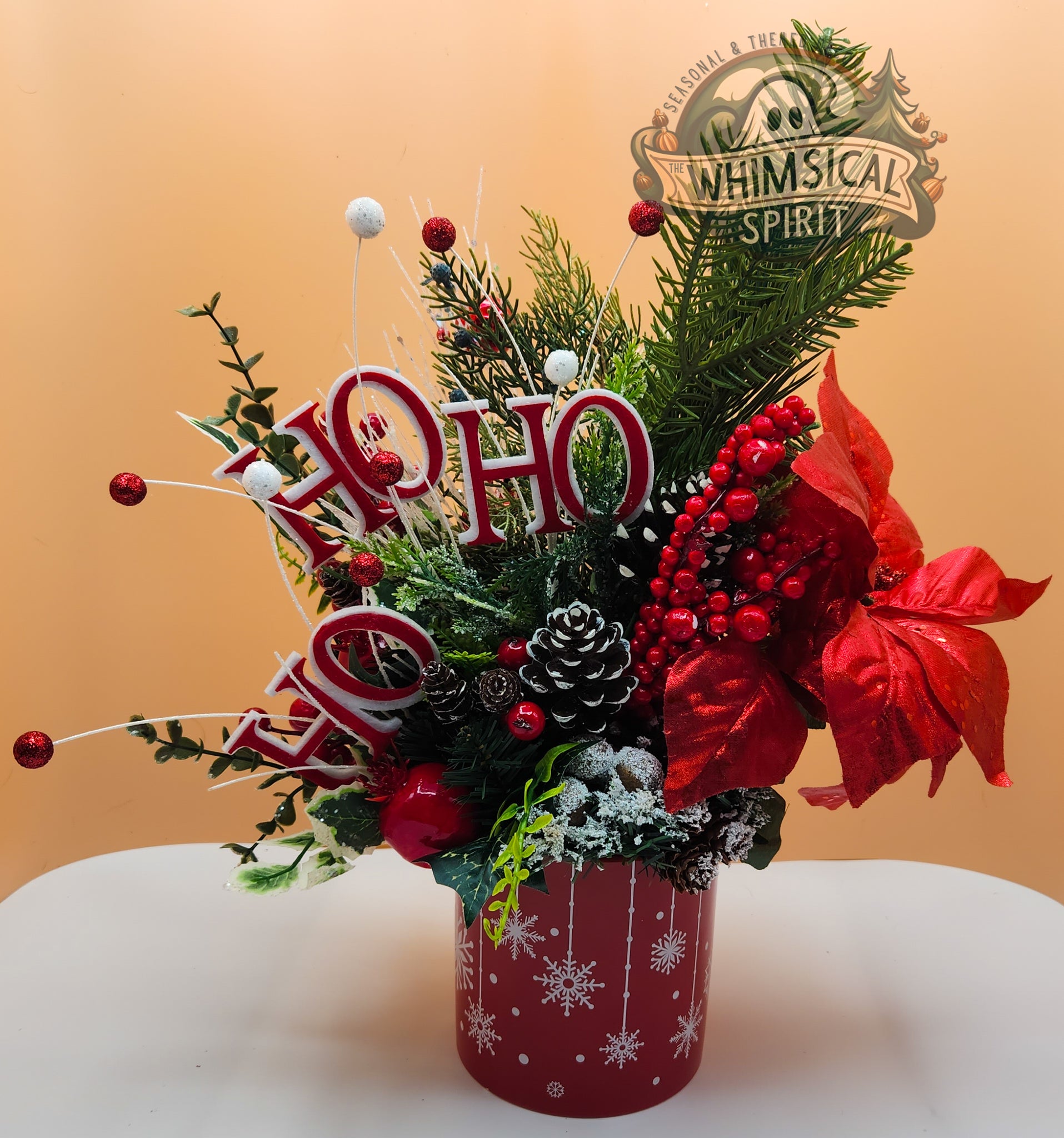 Decorative Christmas arrangement with 'Ho Ho Ho' sign and red vase on a white surface.