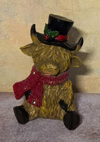 A decorative Highland cow figurine with shaggy golden-brown fur, wearing a black top hat with green holly and a red band, plus a red glittery scarf, seated against a textured beige wall.