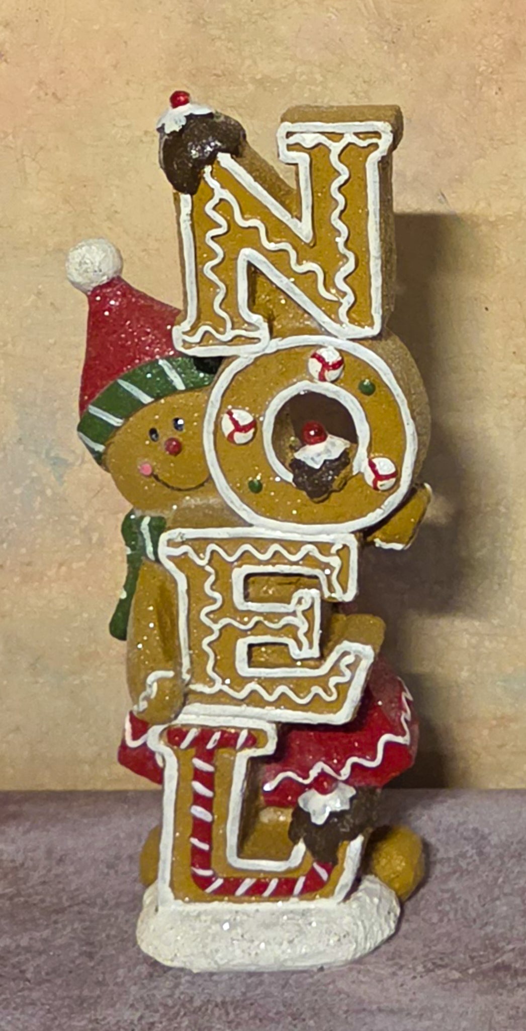A festive gingerbread-themed 'NOEL' decorative sign featuring three illustrated gingerbread cookies spelling out the word, with one cookie wearing a red Santa hat and scarf, set against a neutral beige background.