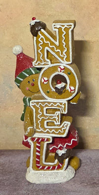 A festive gingerbread-themed 'NOEL' decorative sign featuring three illustrated gingerbread cookies spelling out the word, with one cookie wearing a red Santa hat and scarf, set against a neutral beige background.