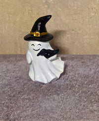 A small Halloween ghost figurine with a white ghost body, black pointed witch hat, and a black bird on its back, set against a textured beige and gray background.