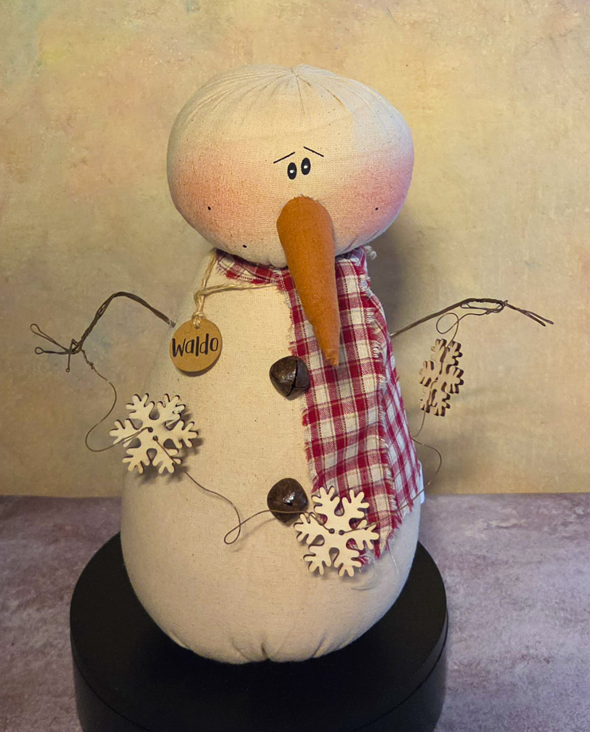 Front view of Waldo snowman soft sculpture with a long carrot nose, red plaid scarf, twig arms, jingle bell buttons, and snowflake garland.