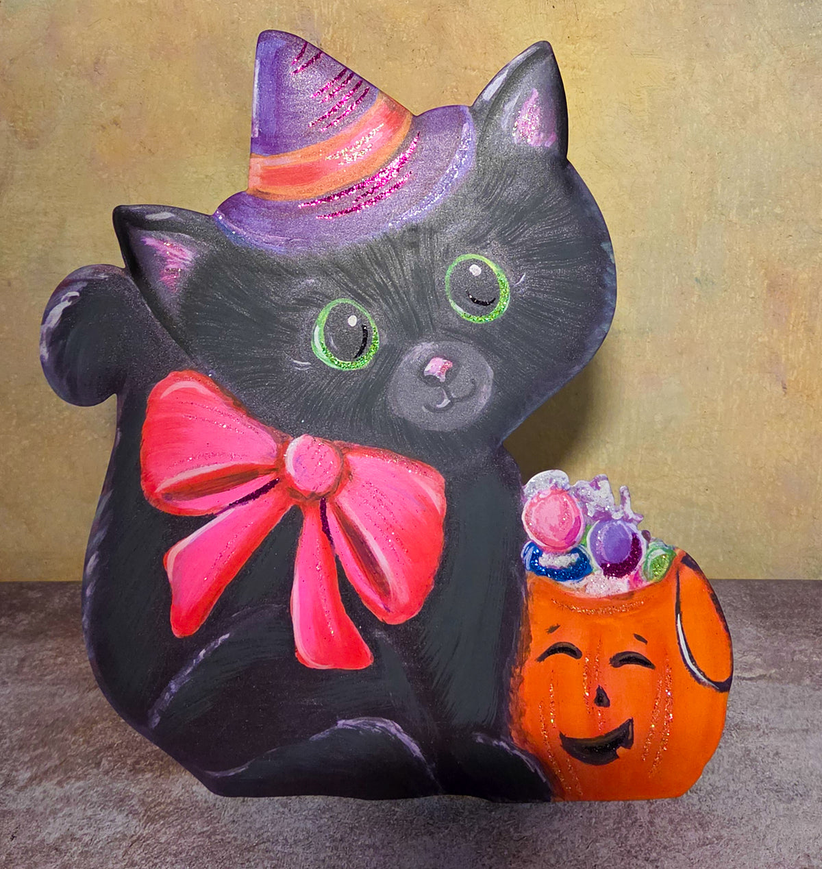Front view of a black cat metal decoration with green eyes, a purple witch hat, pink bow, and orange pumpkin bucket filled with candy.