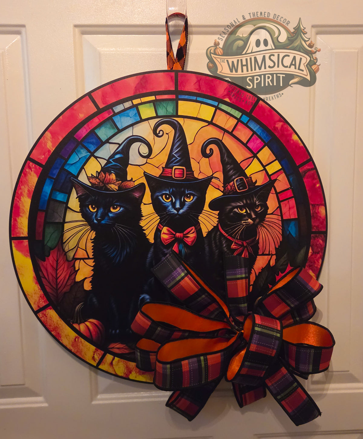 Round Halloween door hanger featuring three black cats in witch hats with a stained glass style background and a large orange and plaid bow, hanging on a white door.