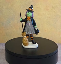 Small Halloween witch resin figurine with green skin, purple hair, pointed black hat, orange bow, gray dress, striped stockings, and broom.