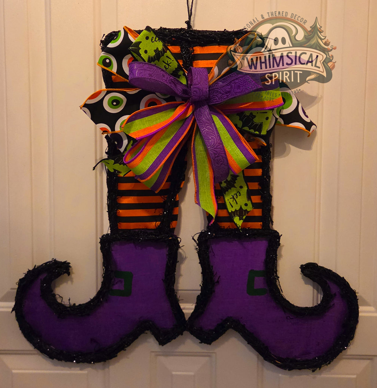 Handcrafted grapevine witch legs Halloween door hanger featuring purple curled boots, orange-and-black striped stockings, and a large purple, green, orange, and black patterned bow.