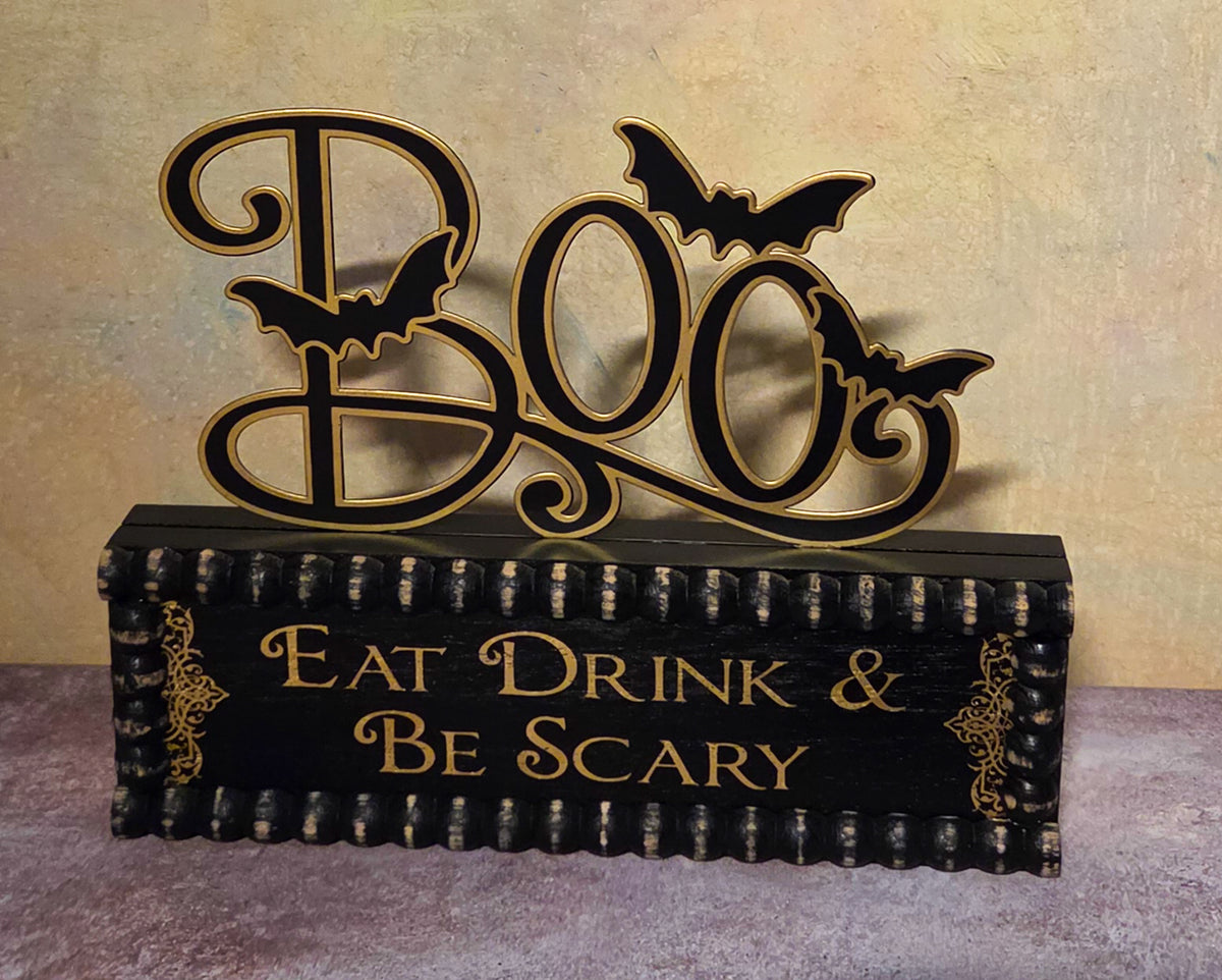 A Halloween-themed tabletop sign with a black base and a large decorative 'BOO' cutout above it, accented with four black bats. The front of the sign reads 'EAT DRINK & BE SCARY' in gold lettering.