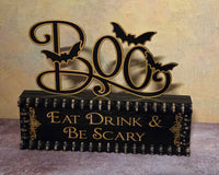 A Halloween-themed tabletop sign with a black base and a large decorative 'BOO' cutout above it, accented with four black bats. The front of the sign reads 'EAT DRINK & BE SCARY' in gold lettering.
