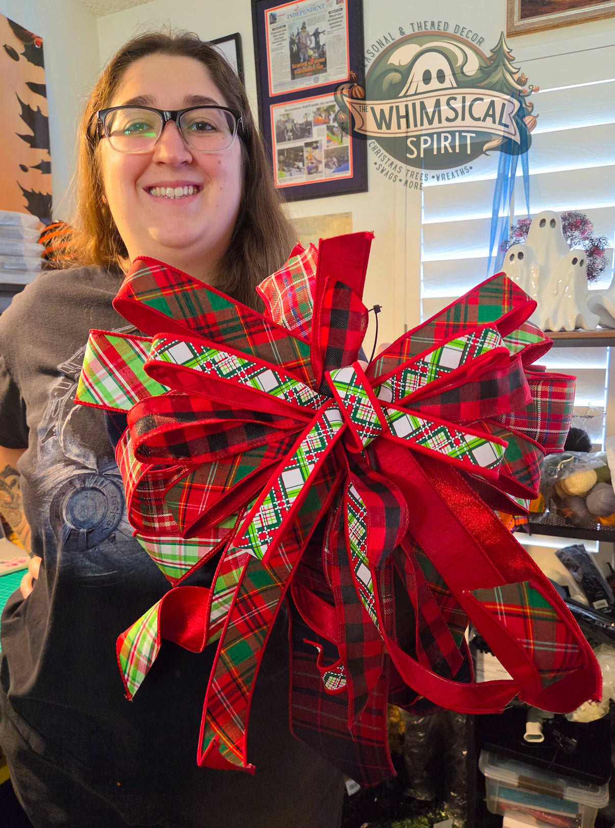 Person standing in a craft room holding a large handmade Christmas bow made with layered red and green plaid ribbon in multiple patterns. The XL bow features a full shape with long ribbon tails and classic Christmas colors.