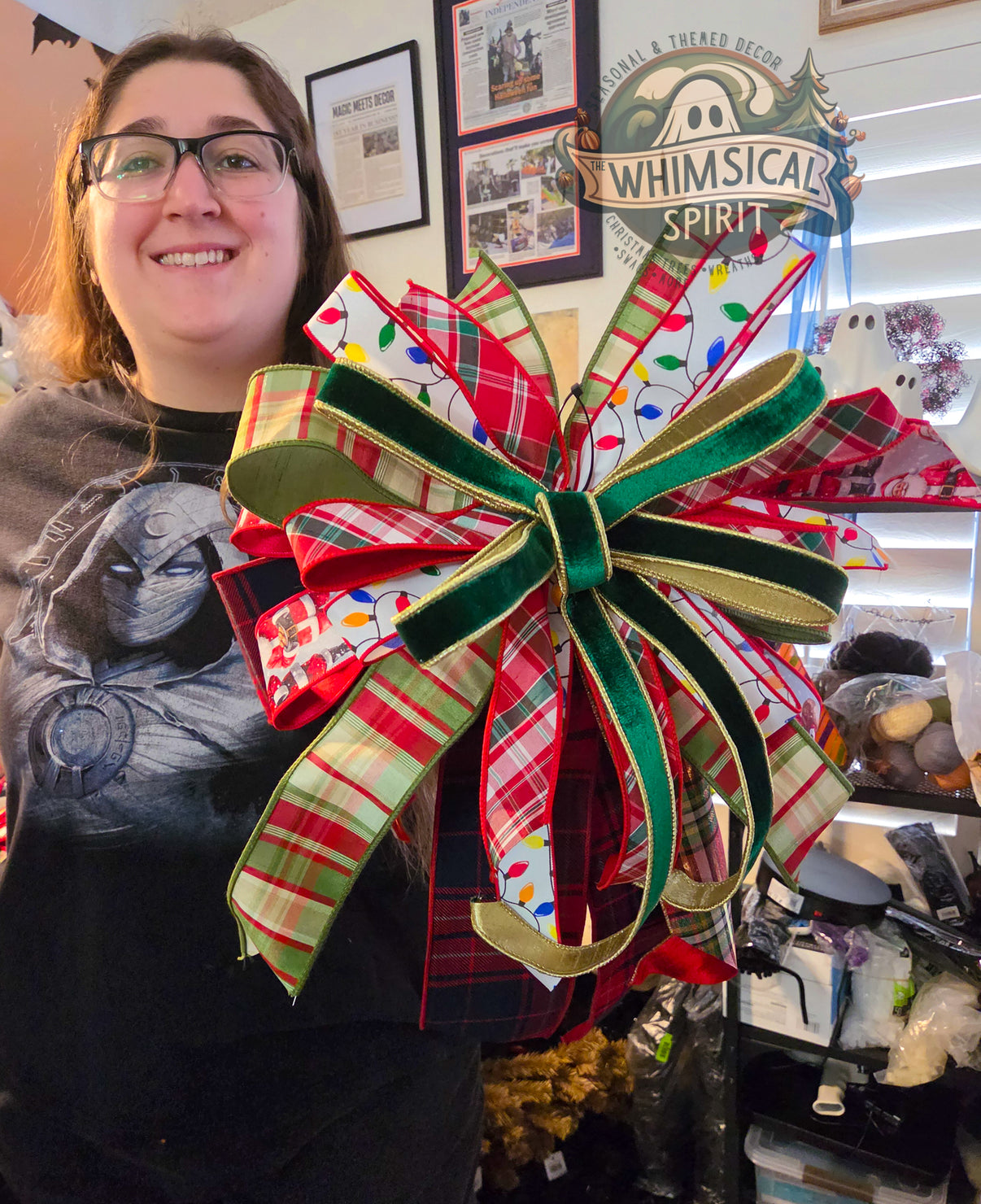 Person standing in a craft room holding a large handmade Christmas bow featuring red and green plaid ribbon, white ribbon with colorful Christmas lights, and green velvet ribbon with gold trim. The XL bow has layered loops and long ribbon tails.