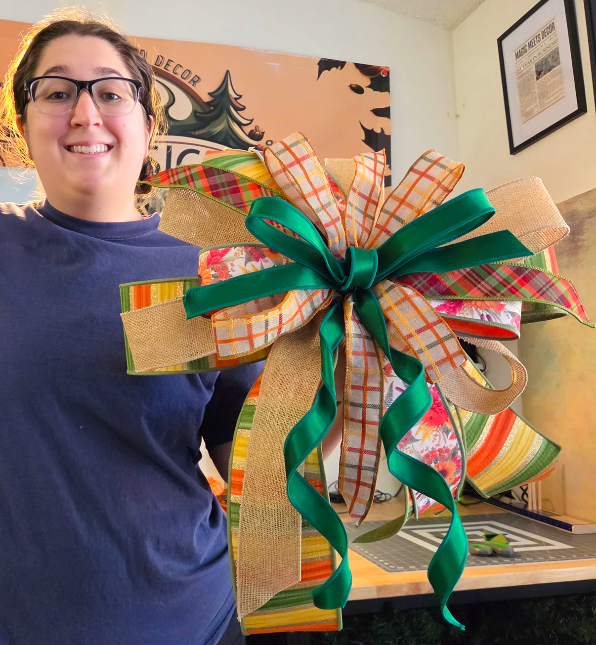 Maker holding an XL fall bow featuring layered green, plaid, burlap, checked, striped, and floral ribbon.