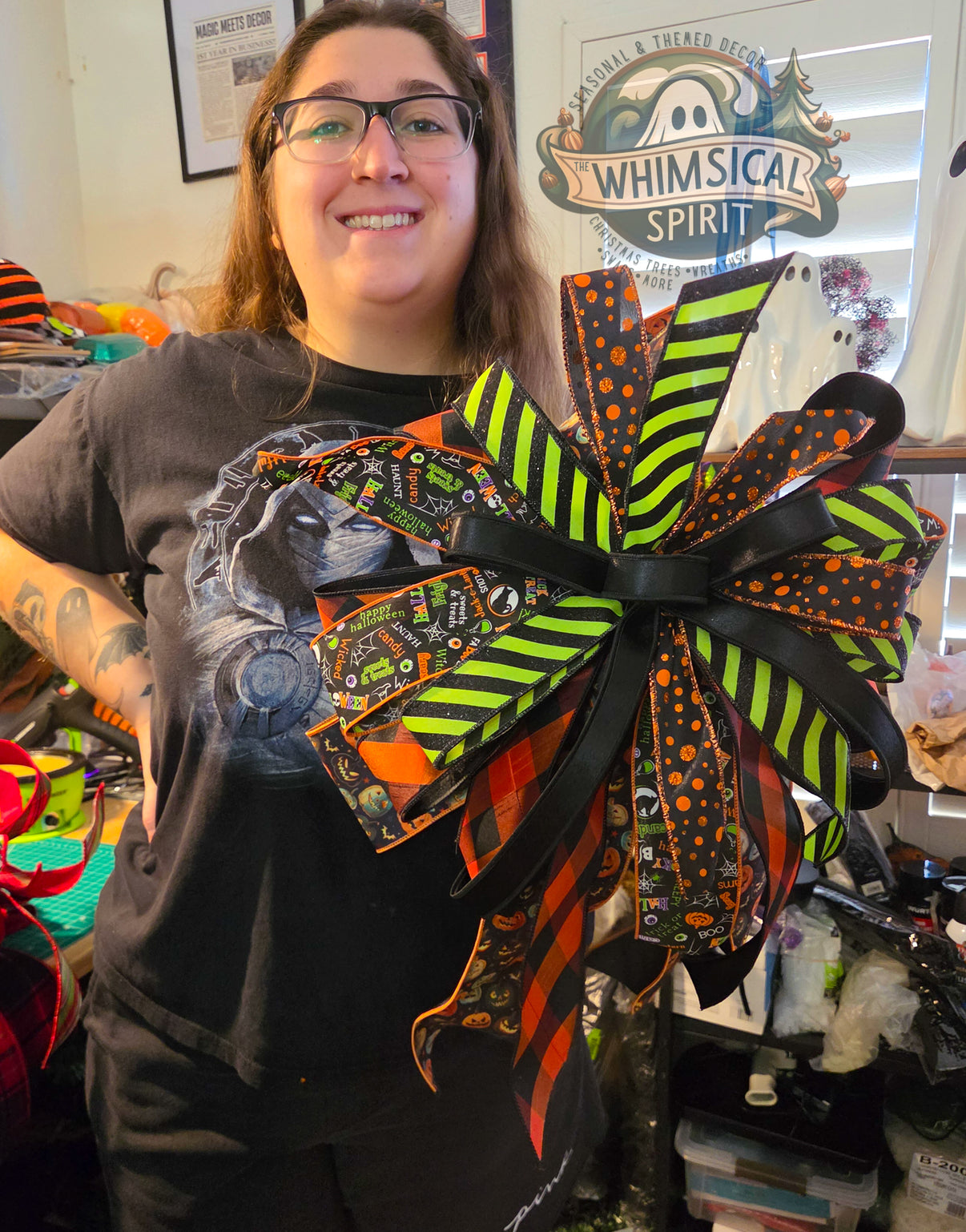 Person standing in a craft room holding a large handmade Halloween bow made with black, orange, and lime green ribbon. The XL bow features striped, polka dot, buffalo check, and Halloween-themed ribbon patterns with long ribbon tails.