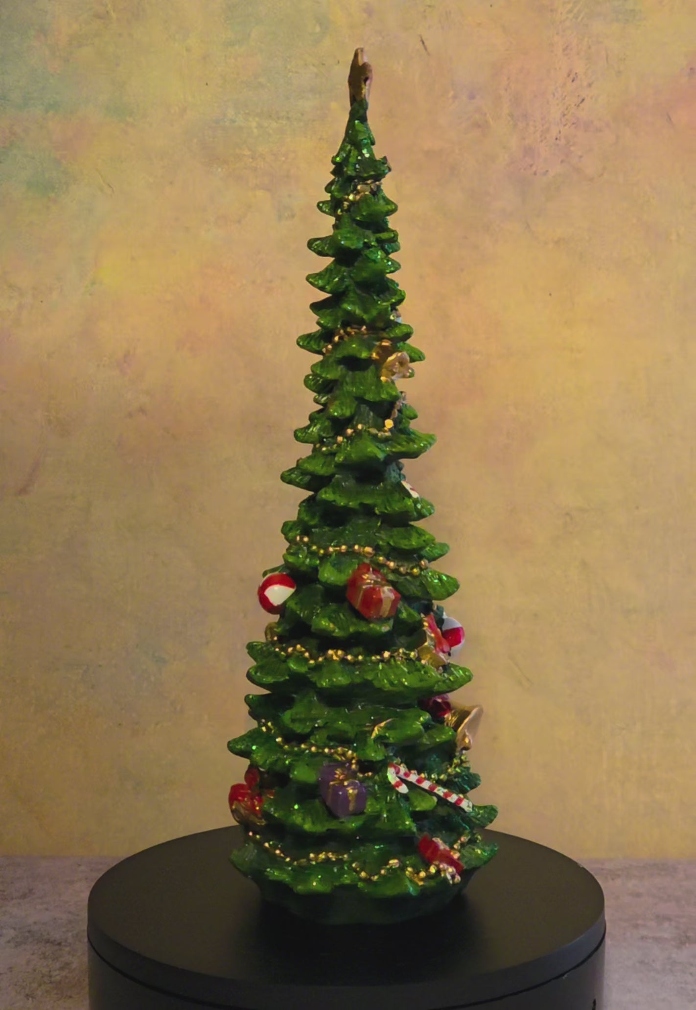 Short turntable video of a decorated resin Christmas tree tabletop figurine with a gold star topper, gold garland, colorful ornaments, bells, and miniature gift accents, rotating to show all sides.