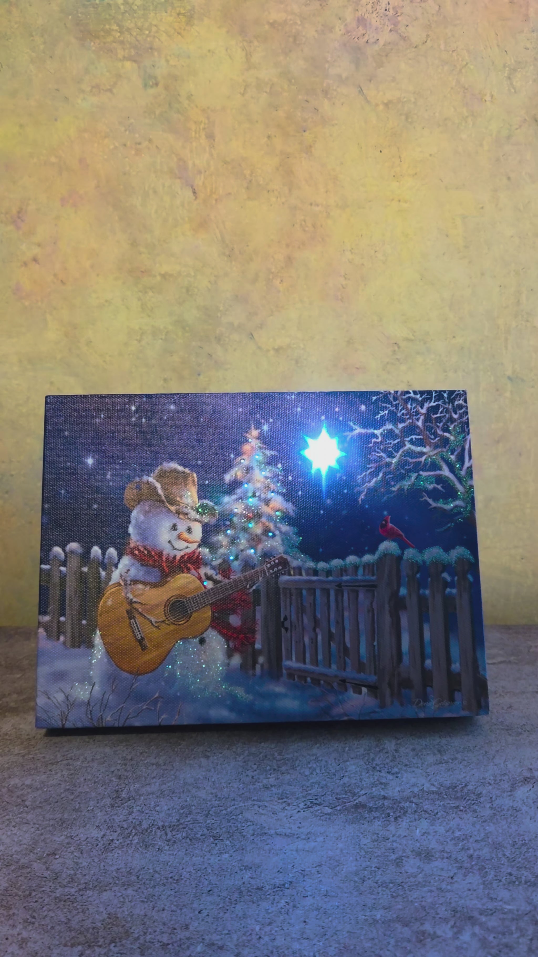 Video showing the light-up function on Guitar Snowman, a Christmas tabletop canvas featuring a cowboy snowman playing guitar beside a glowing tree, snowy fence, red cardinal, and starry winter sky.